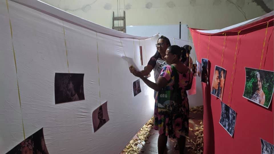 Mulheres observando retratos de pessoas negras pendurados em painéis de tecido branco e vermelho