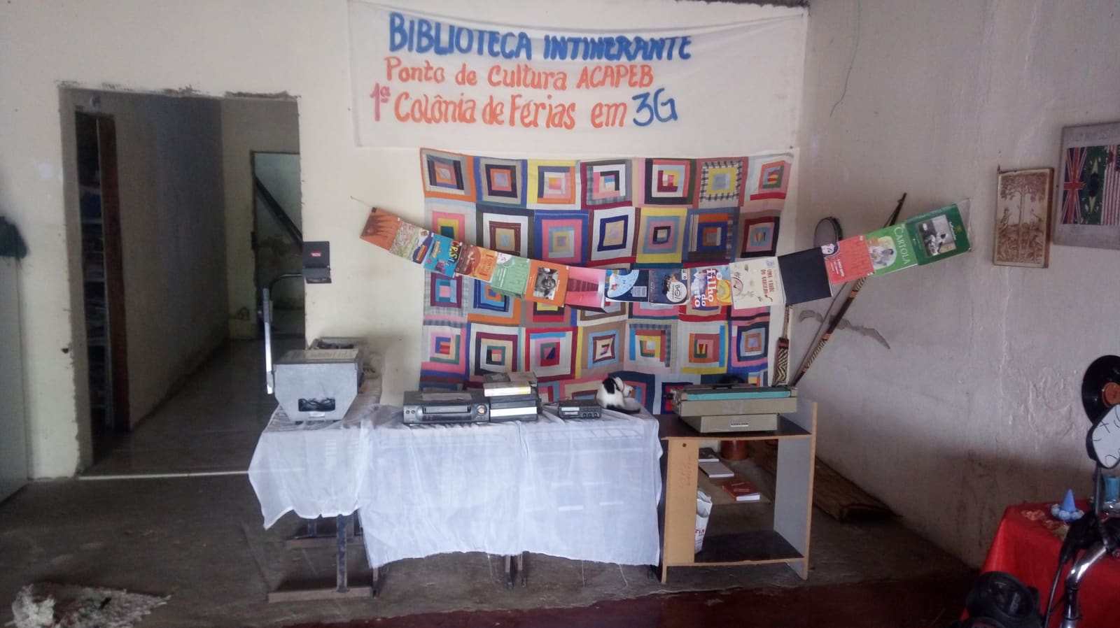 Mesa com equipamentos antigos expostos diante de uma colcha de retalhos e da faixa da Biblioteca Itinerante